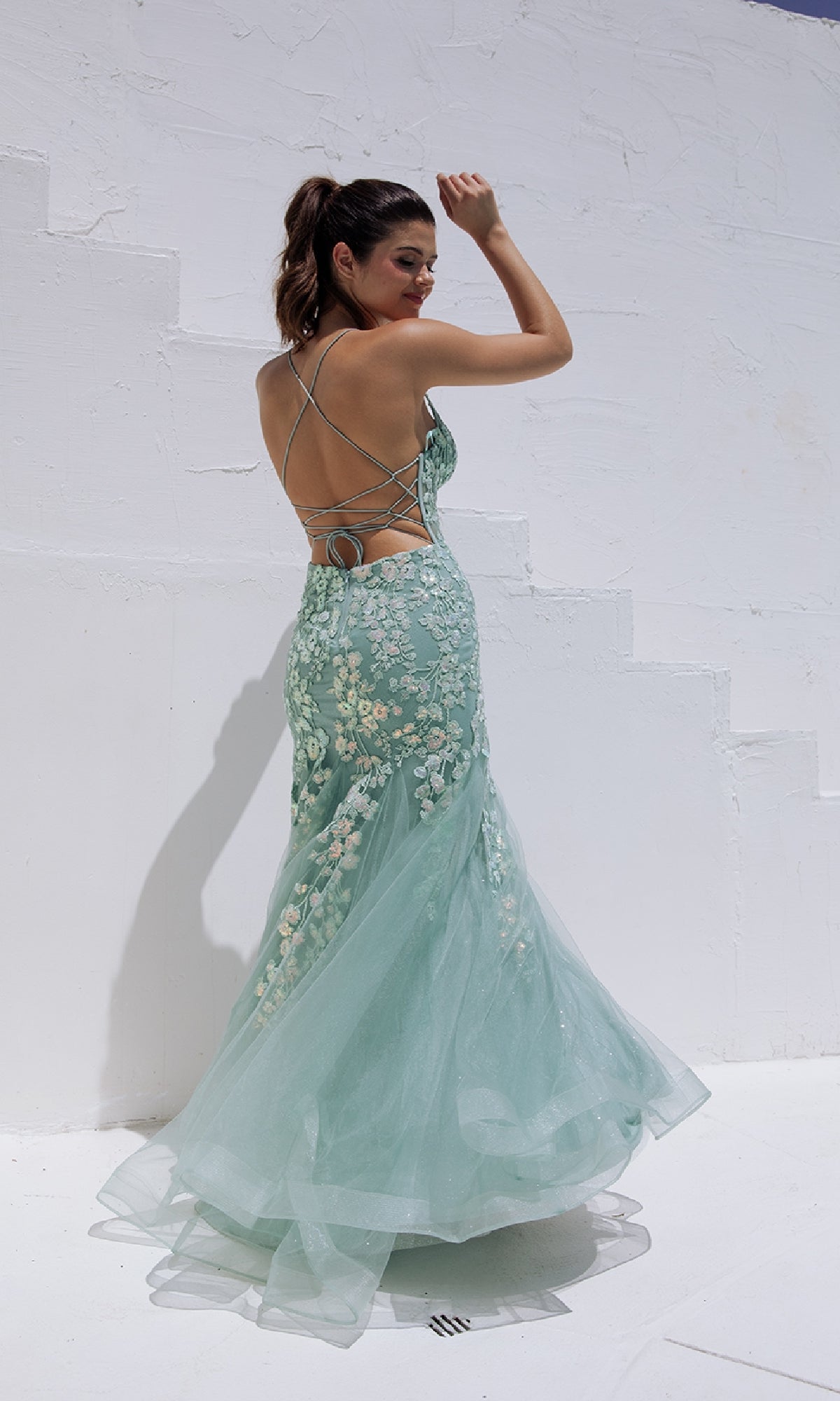 A woman in the Sequin-Floral Long Mermaid Prom Dress: Eureka 9696 poses outdoors against a white textured wall, her hair tied back and one arm raised.